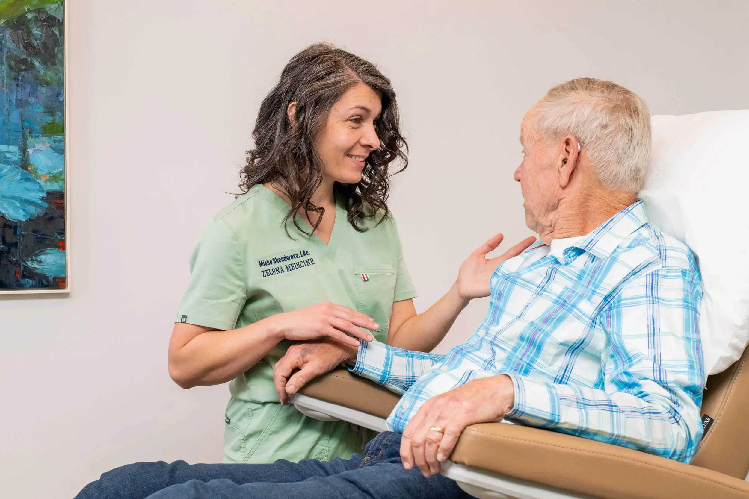 Mishe Skenderova, acupuncturist at Zelena Medicine in Basalt, Colorado talking to an elderly male patient who is sitting in a medical examination chair.