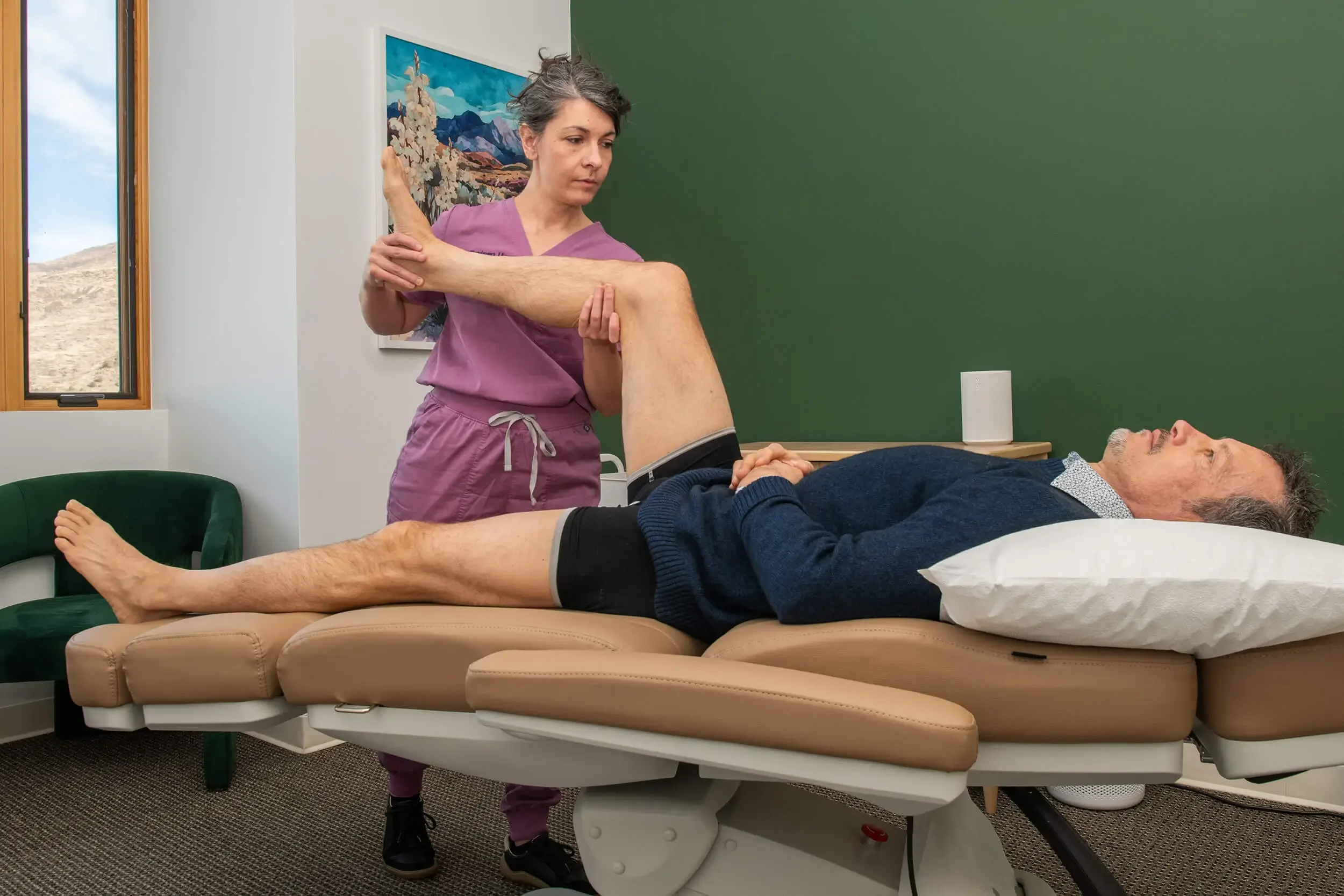 Mishe Skenderova, acupuncturist at Zelena Medicine in Basalt, Colorado assisting a patient with leg therapy on an examination table in a medical clinic.