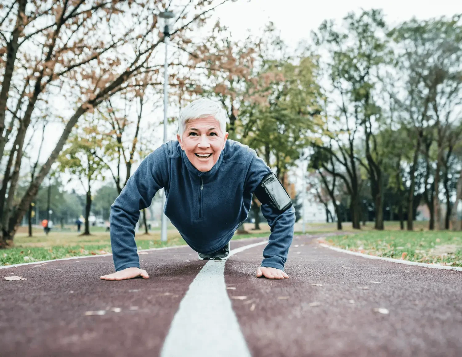 An older woman with short white hair in a blue athletic jacket doing a push-up on a running track after women's holistic health visit with Zelena Medicine.