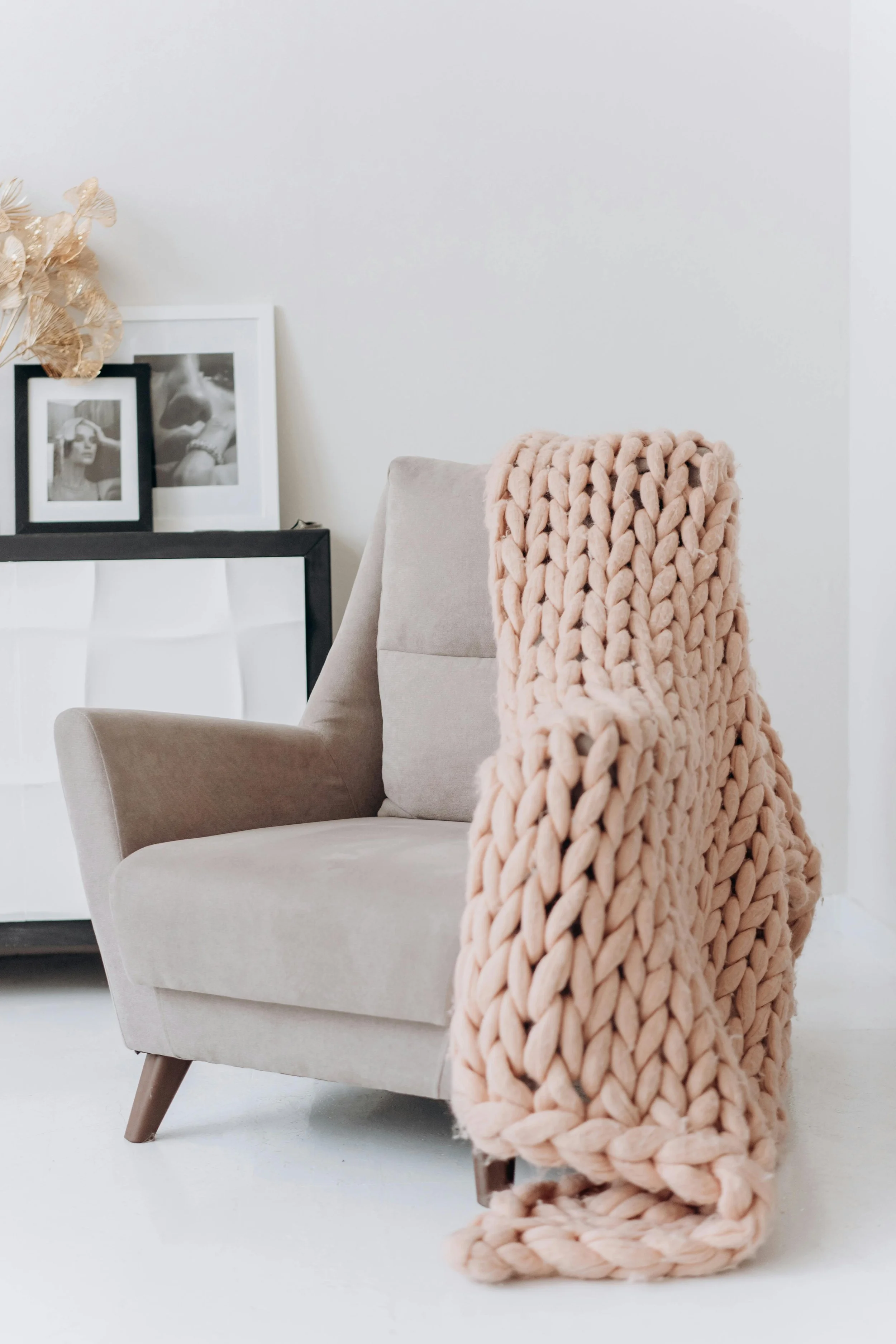A cozy beige armchair with a chunky blush pink knit blanket draped over one arm, situated in a minimalist white room with framed black and white photographs and dried floral decor in the background.