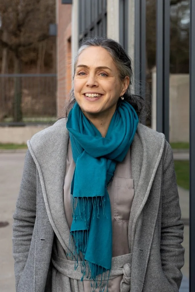 Foto von Anne Schuester. Lächelnden Frau mit grauen, nach hinten gebundenen Haaren. Sie trägt einen blauen Schal und einen grauen Mantel.