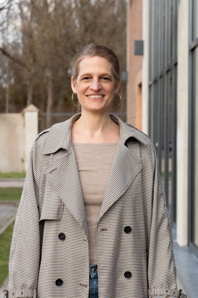 Foto von Daniela Aue. Lächelnde Frau mit kurzen Haaren und großen goldenen Ohrringen. Sie trägt einen schwarz-weißen Mantel.