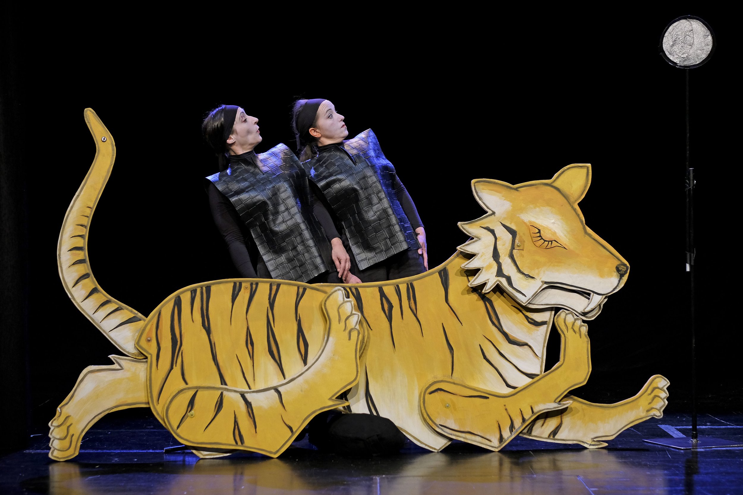 Zwei Performerinnen in schwarzen Kostümen stehen hinter einer großen, stilisierten Tigerfigur auf einer Bühne. Sie lehnen sich zur Seite und blicken gemeinsam nach oben, vor schwarzem Hintergrund. 