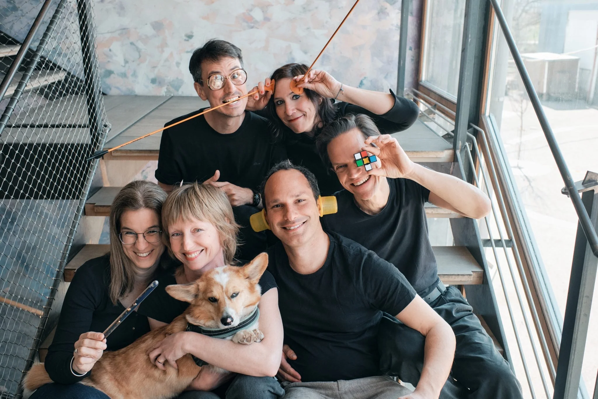 Sechs Personen sitzen gemeinsam auf einer Treppe und lachen in die Kamera. Einige halten kleine Gegenstände, ein Hund liegt entspannt auf dem Schoß.