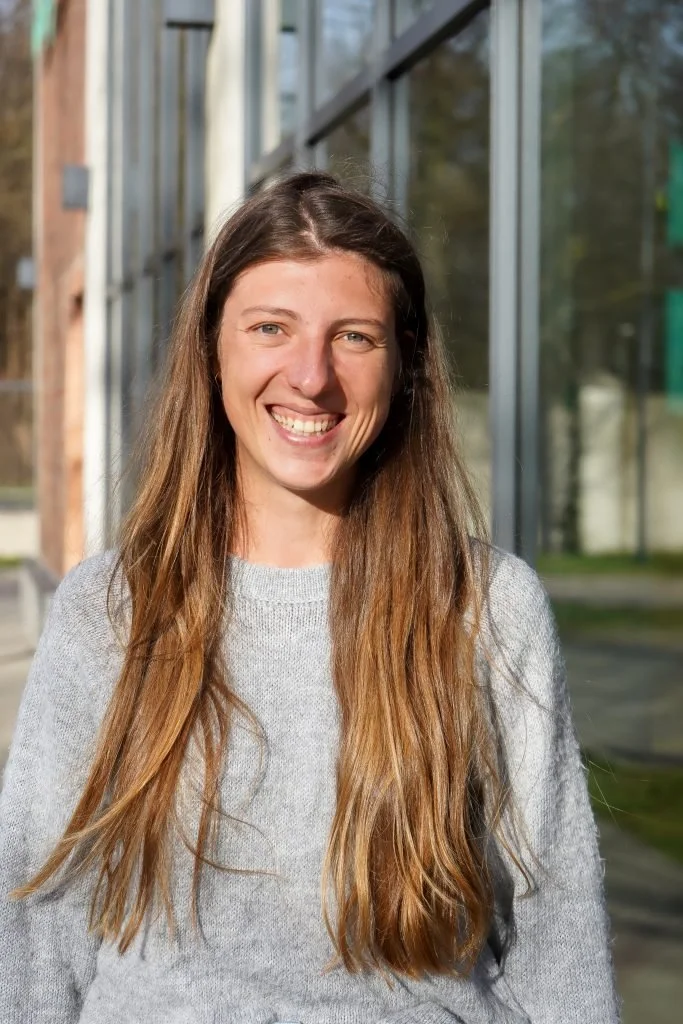 Foto von Paulina Platzer. Lächelnde junge Frau mit langen braunen Haaren. Sie trägt einen grauen Pullover.