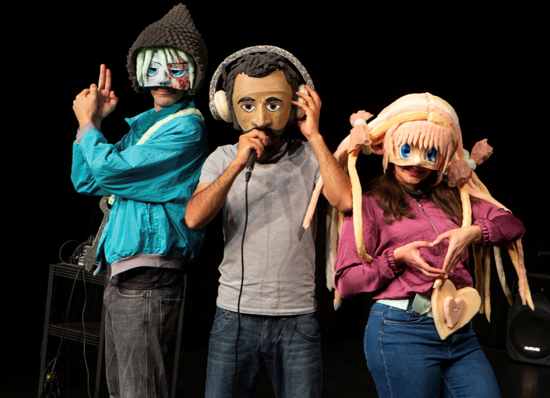 Drei Personen tragen große, fantasievolle Masken. Eine hält ein Mikrofon. Sie erinnern an Figuren aus einem Comic. Dunkler Bühnenhintergrund.