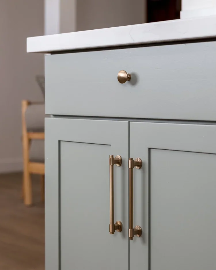 Close-up of a light gray kitchen cabinet with gold handles and a drawer with a round gold knob.