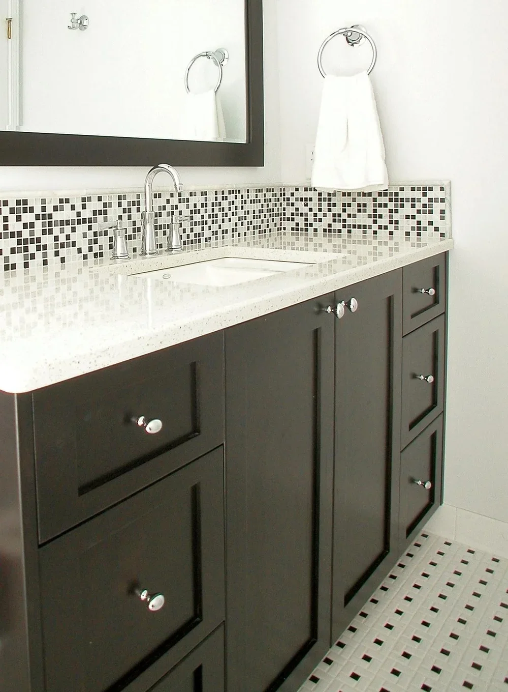 A bathroom vanity with a black cabinet, a white speckled countertop, a mosaic tile backsplash, and a large mirror. There is a towel ring with a white towel and a chrome faucet.