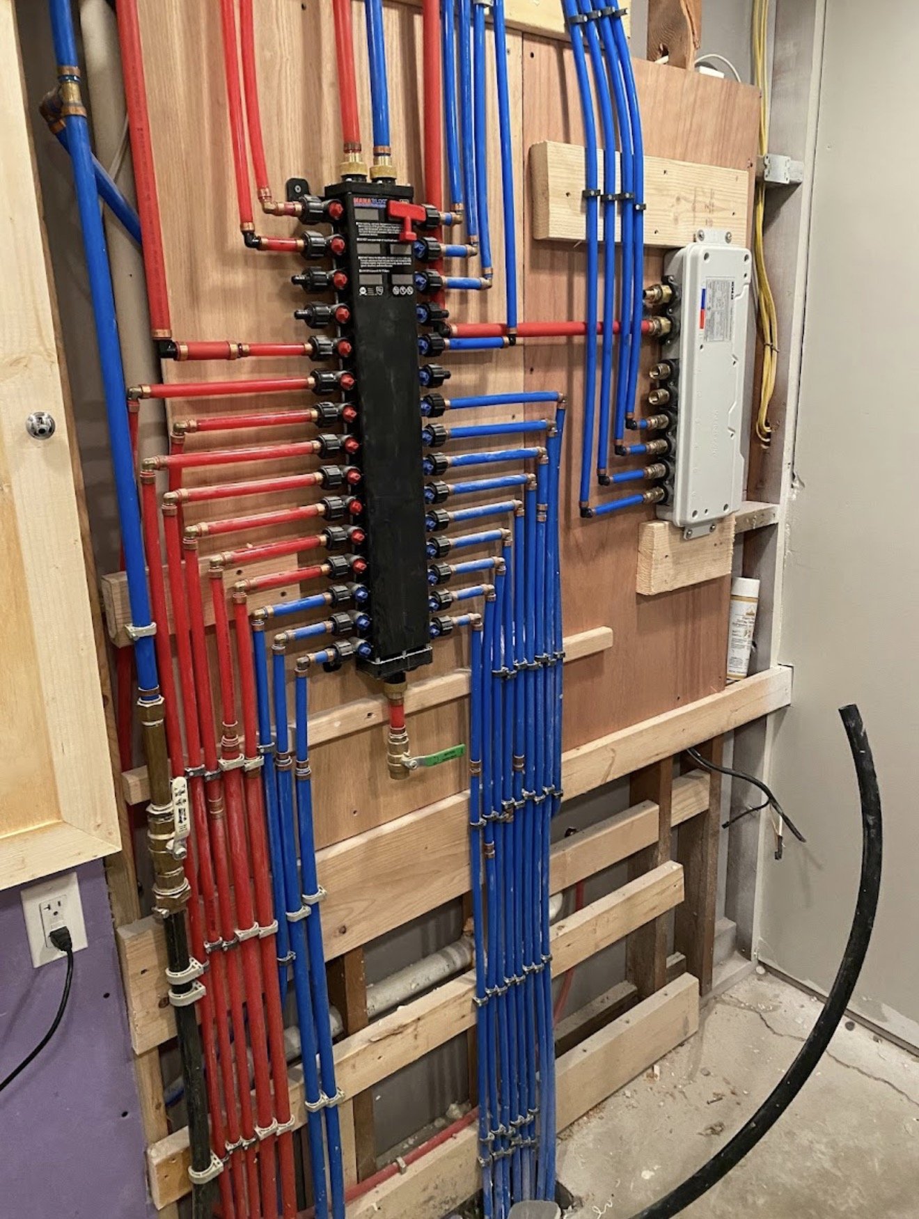 A plumbing manifold with red and blue PEX pipes connected, mounted on a wooden wall in a construction or renovation setting.