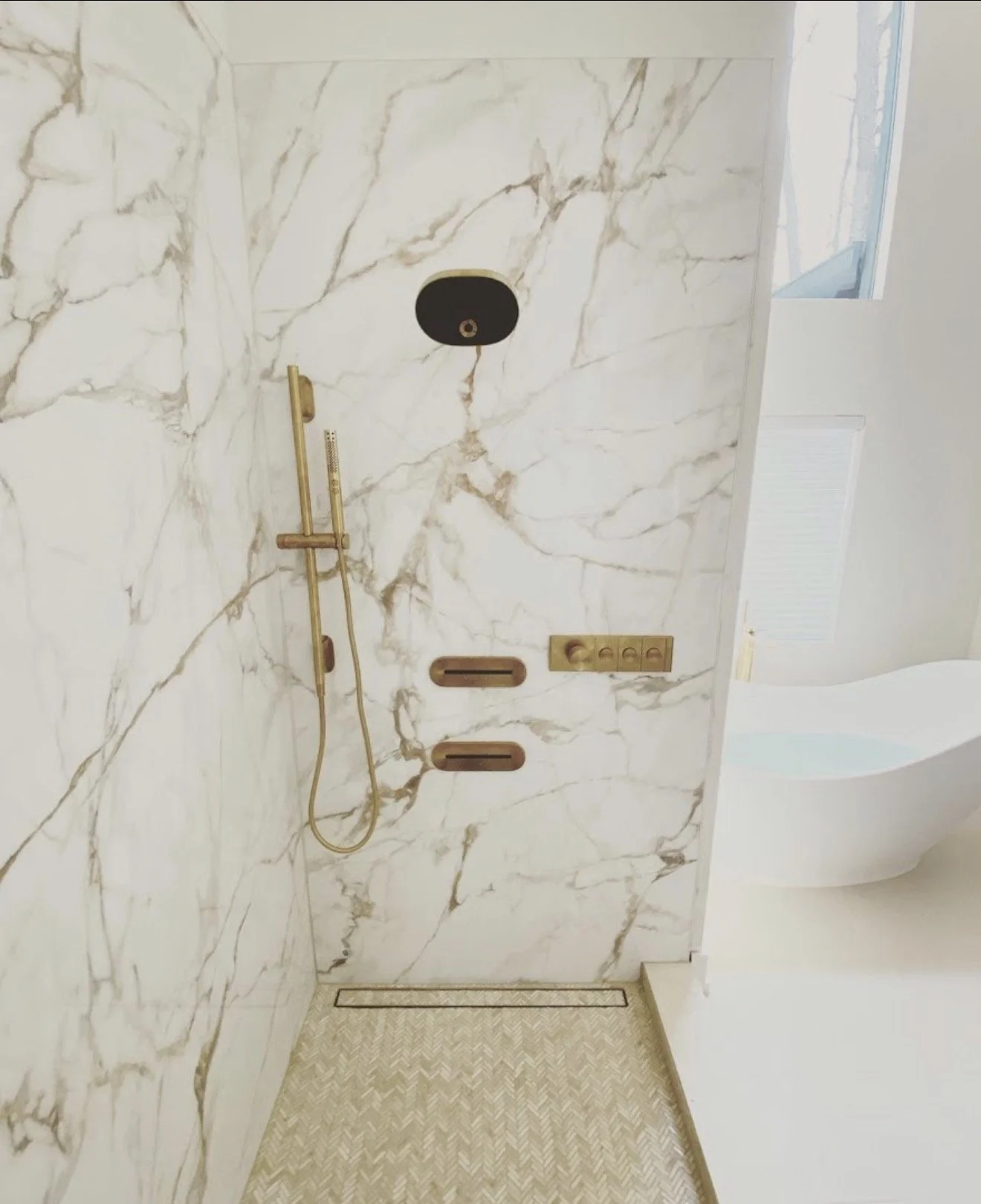 Modern shower with marble walls, gold fixtures, and built-in shelves, adjacent to a white bathtub and window with natural light.