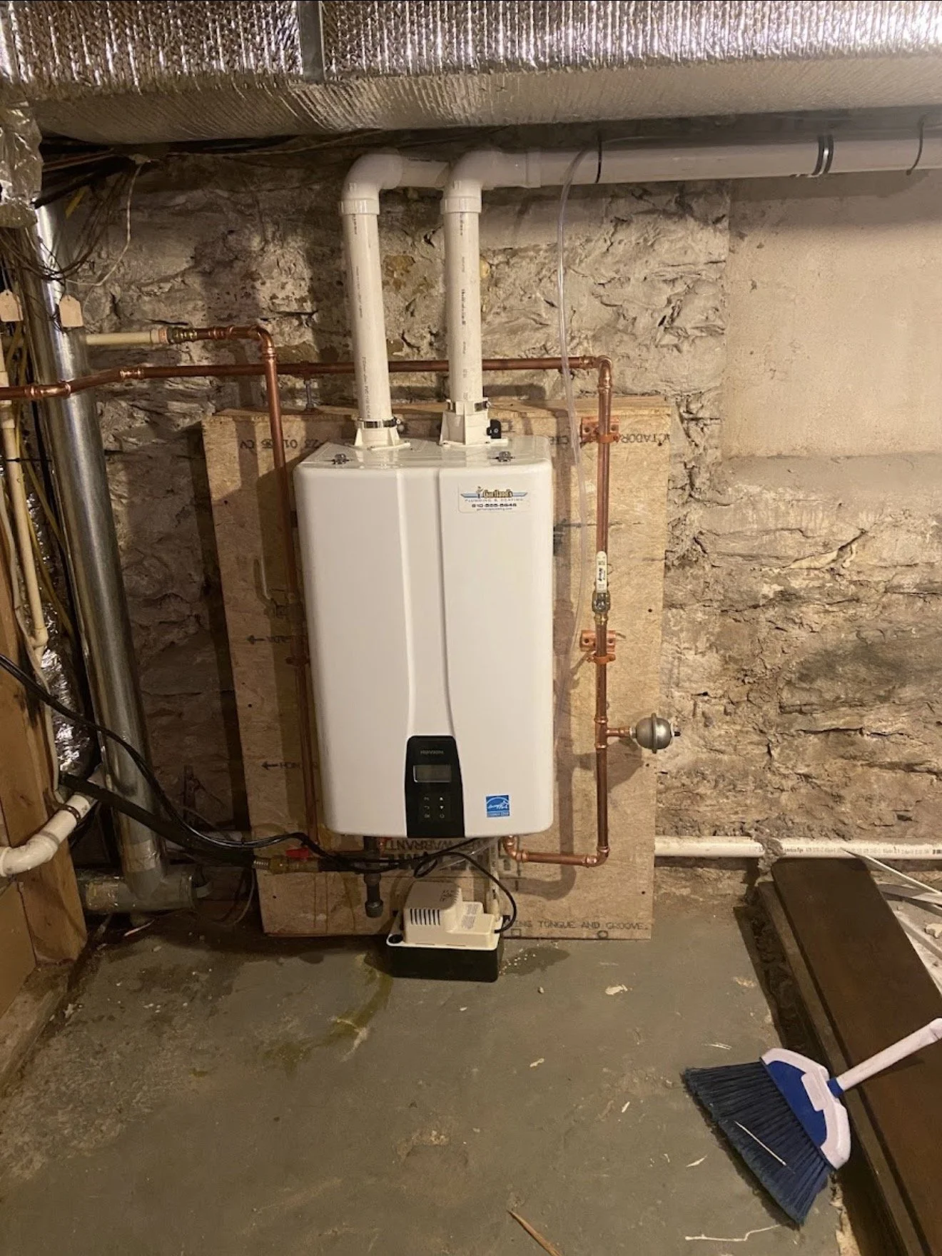 A furnace with a digital display mounted on a concrete floor in a basement. Copper pipes connect to the furnace, and a gray wall with stone and drywall surrounds it. A broom leans against a cabinet to the right.
