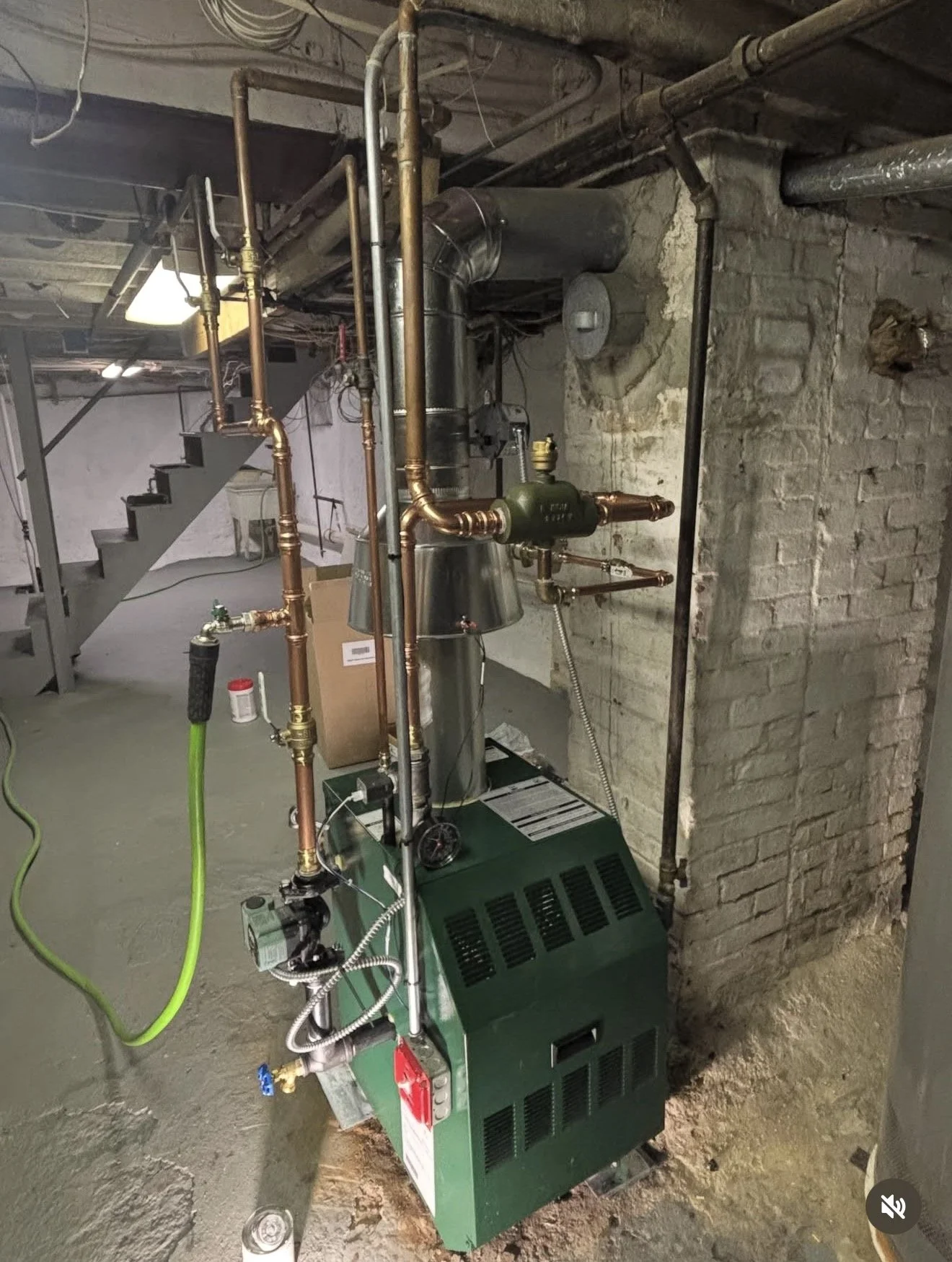 Industrial boiler system with copper pipes, a green control box, and a metal duct in a basement near brick walls and concrete floor.