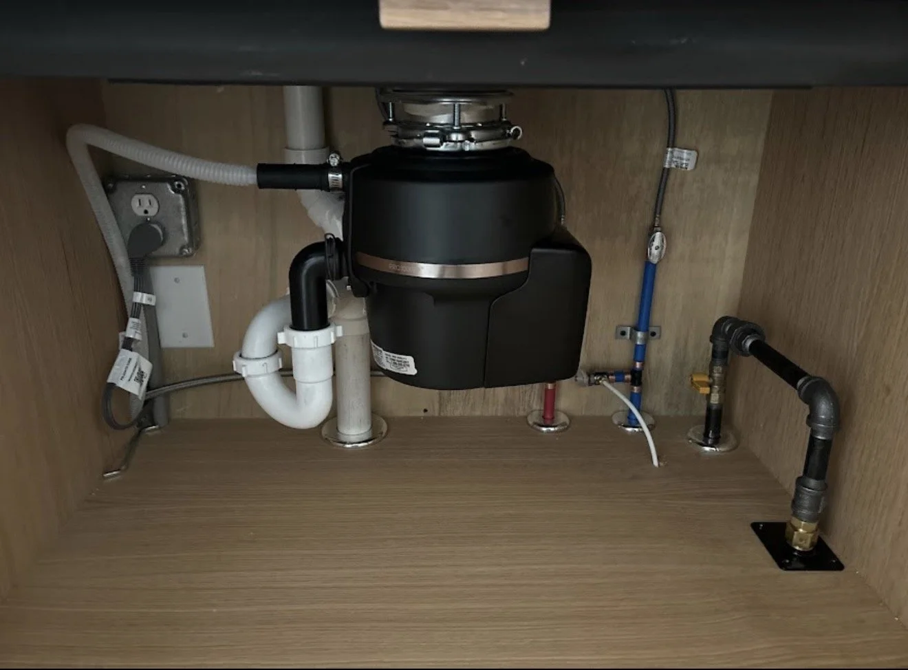 Plumbing under a kitchen sink with a garbage disposal unit, various pipes, and electrical wiring visible inside a wooden cabinet