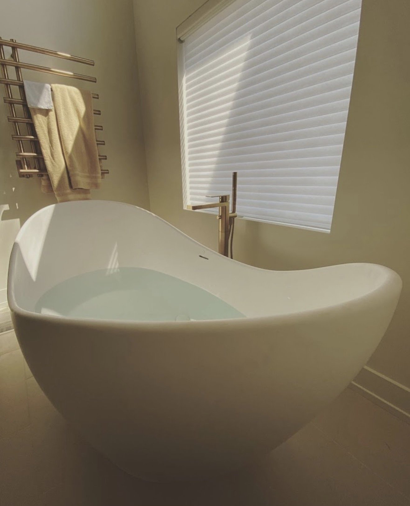 Modern bathtub filled with water in a bathroom with a window and beige towels on a towel rack.