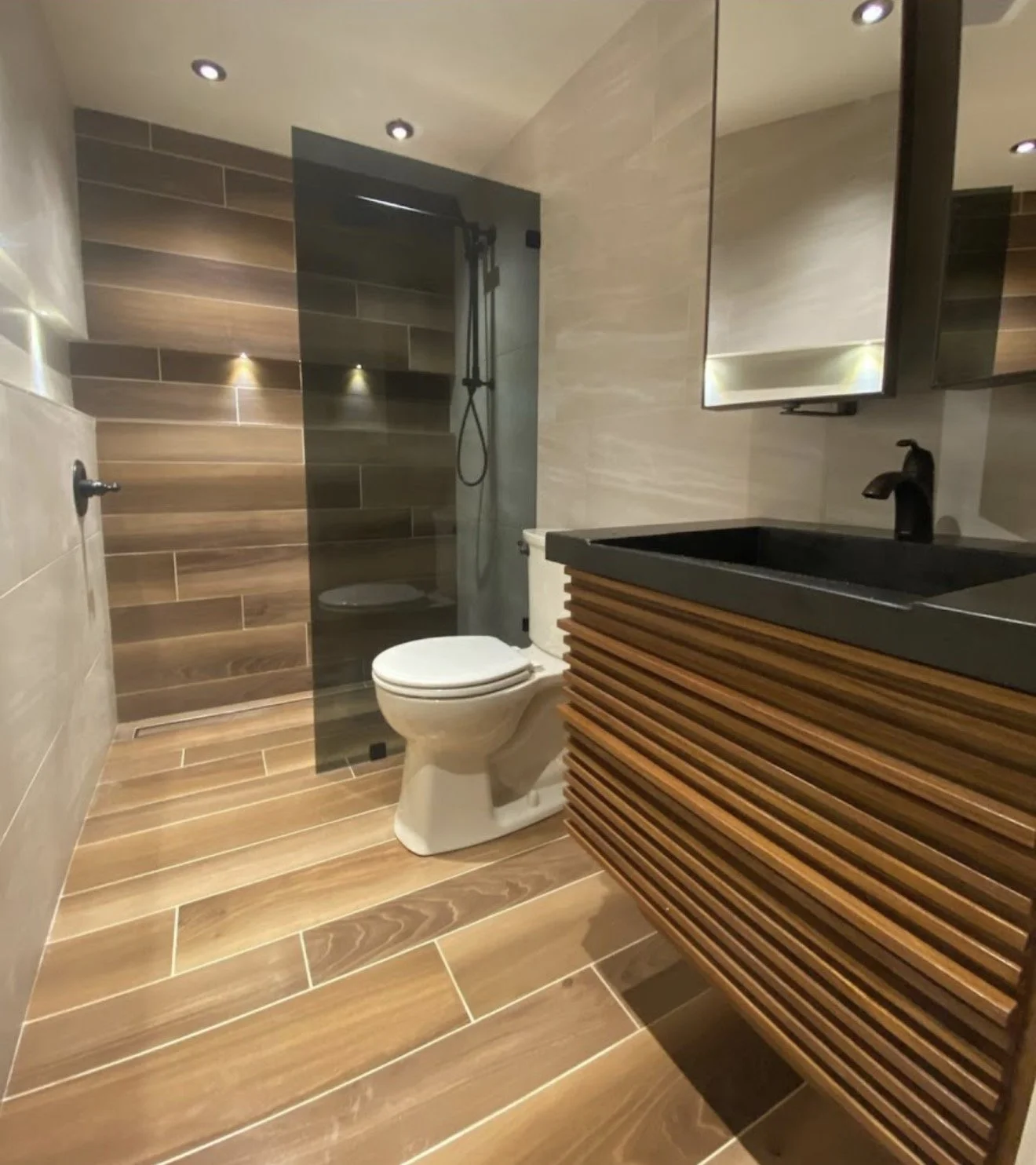 Modern bathroom with wooden flooring, a toilet, a black countertop basin, a mirror, and a walk-in shower with a glass door.