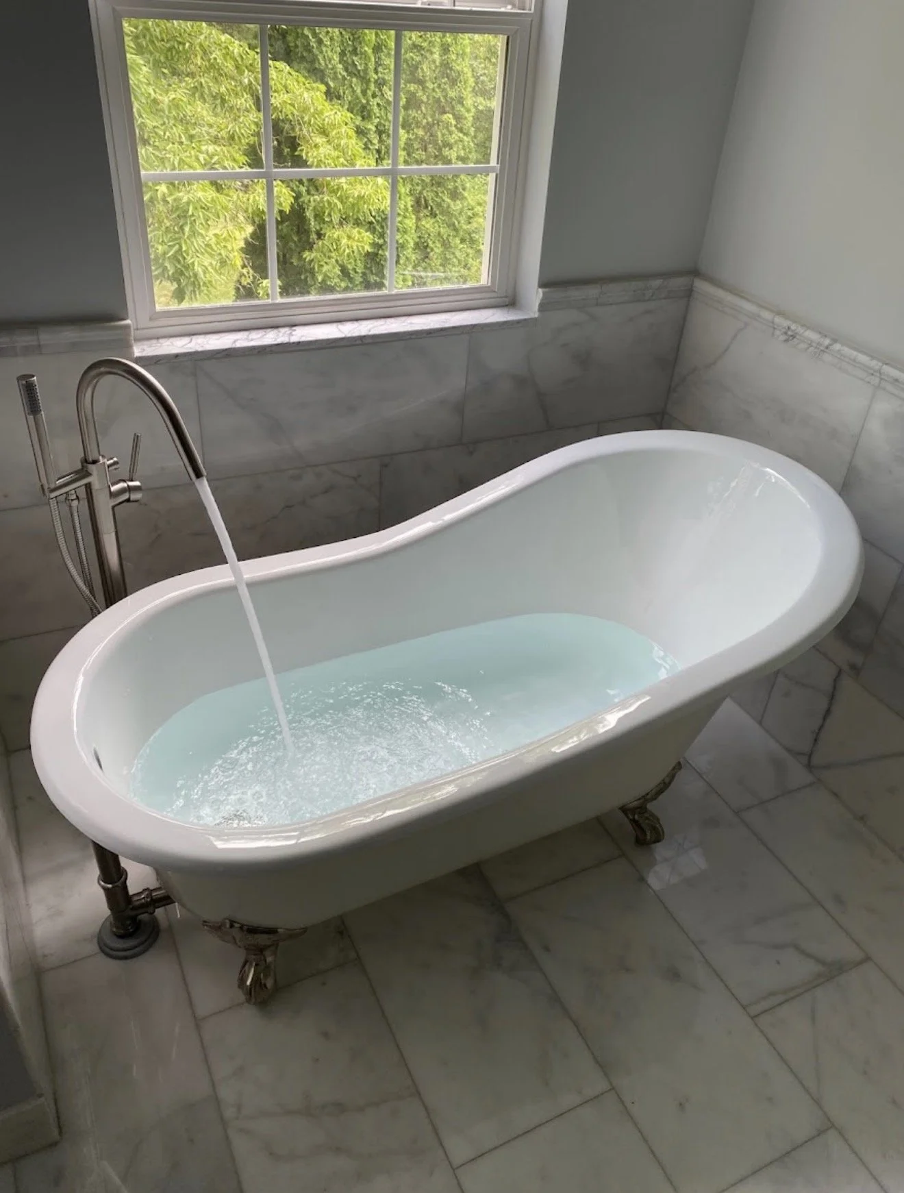 A vintage-style white bathtub with clawfoot legs filled with water, positioned in front of a window showing green trees outside. The bathtub has a silver faucet with water running into it, and the room has marble tile flooring and walls.