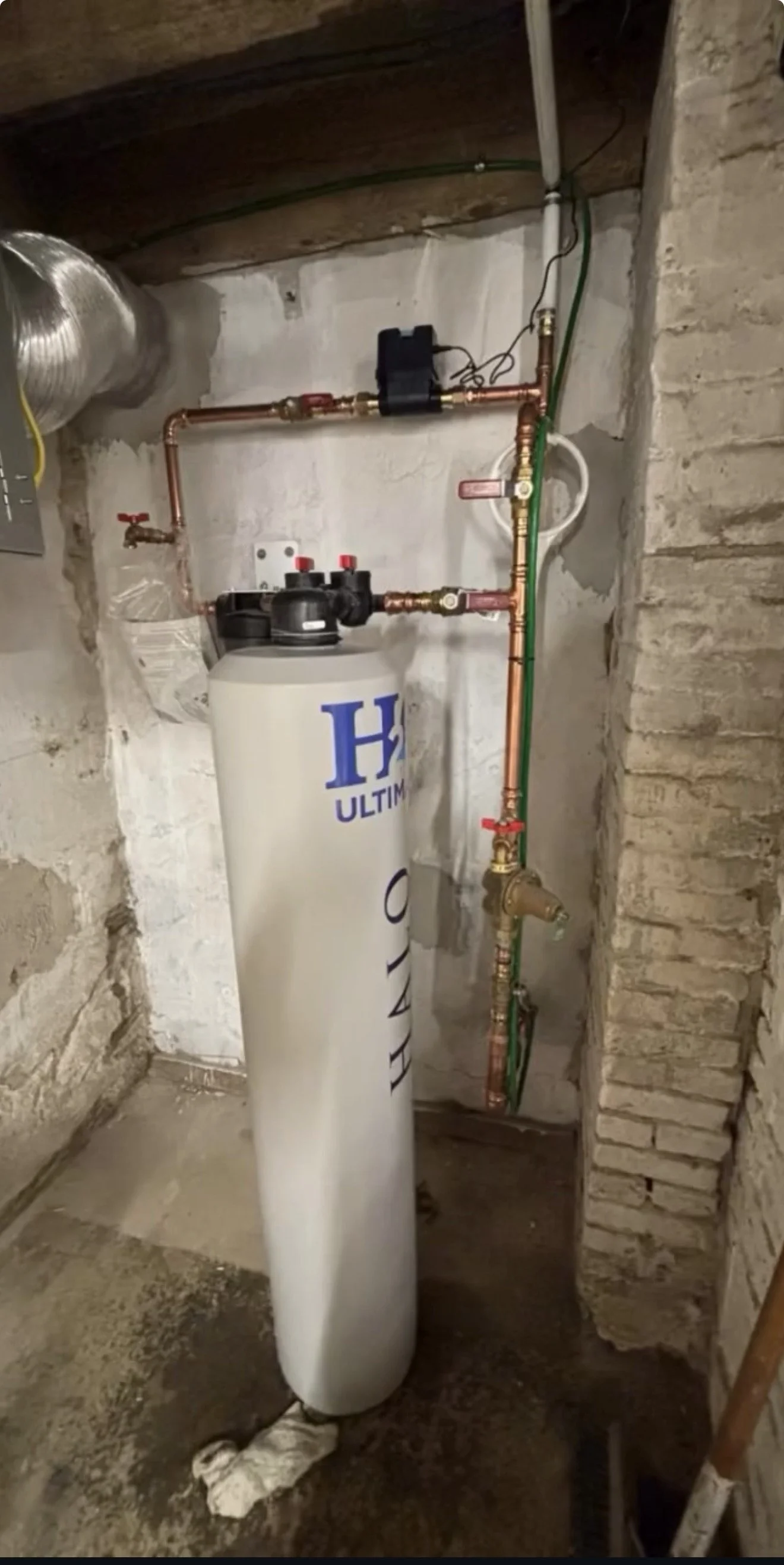 Water softener system with copper pipes, valves, and control unit in a basement utility area.