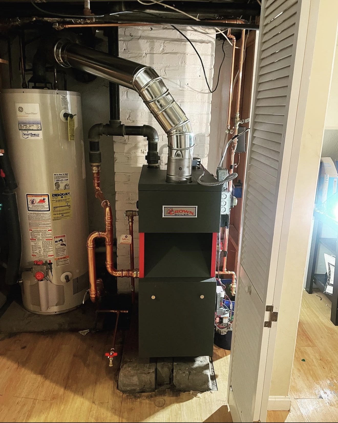 A boiler room showing a boiler labeled Crown, connected to copper and black pipes, next to a water heater, with a brick wall and a wooden floor.