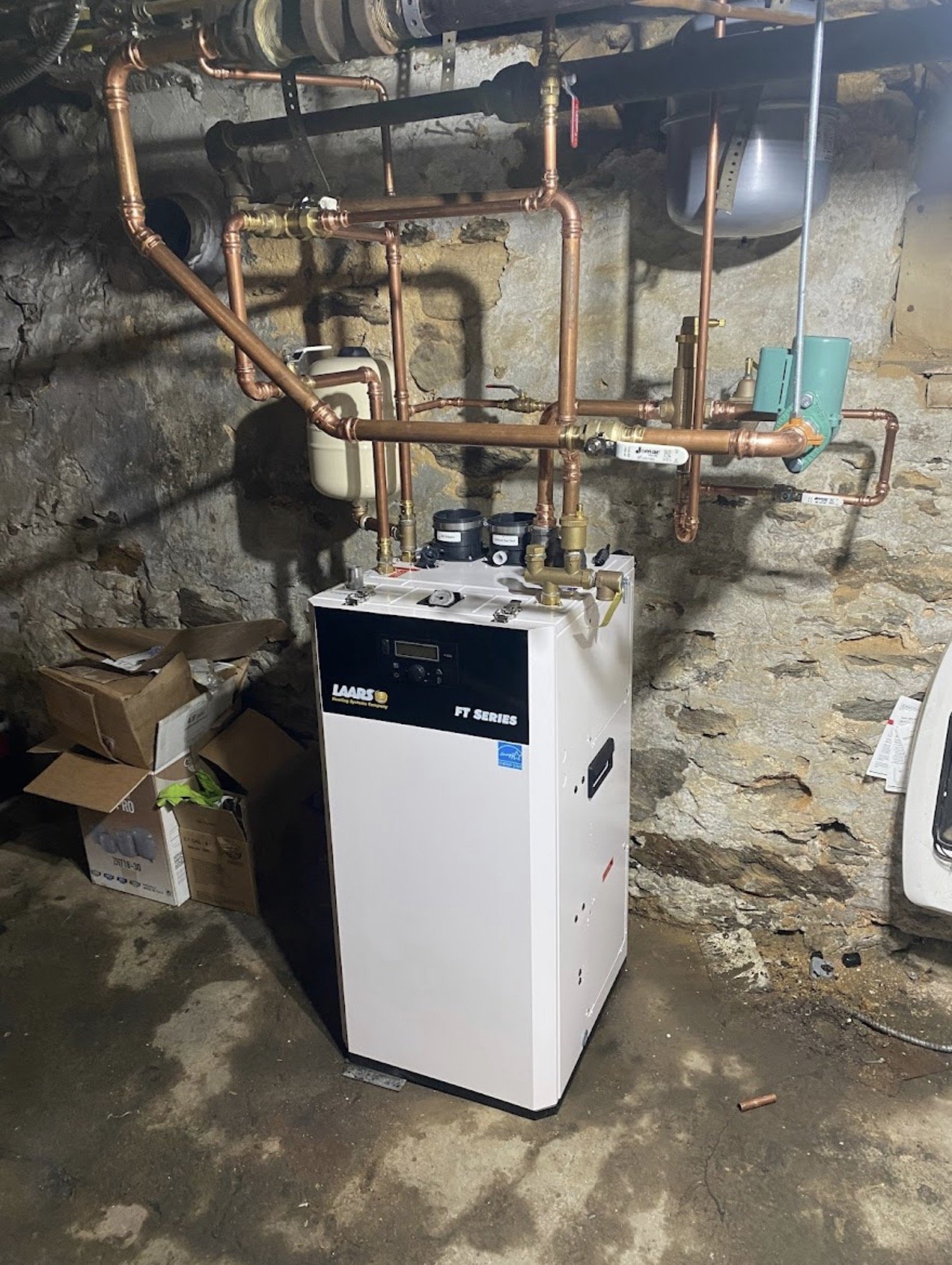 A boiler system with copper pipes mounted on a stone wall in a basement or utility room.