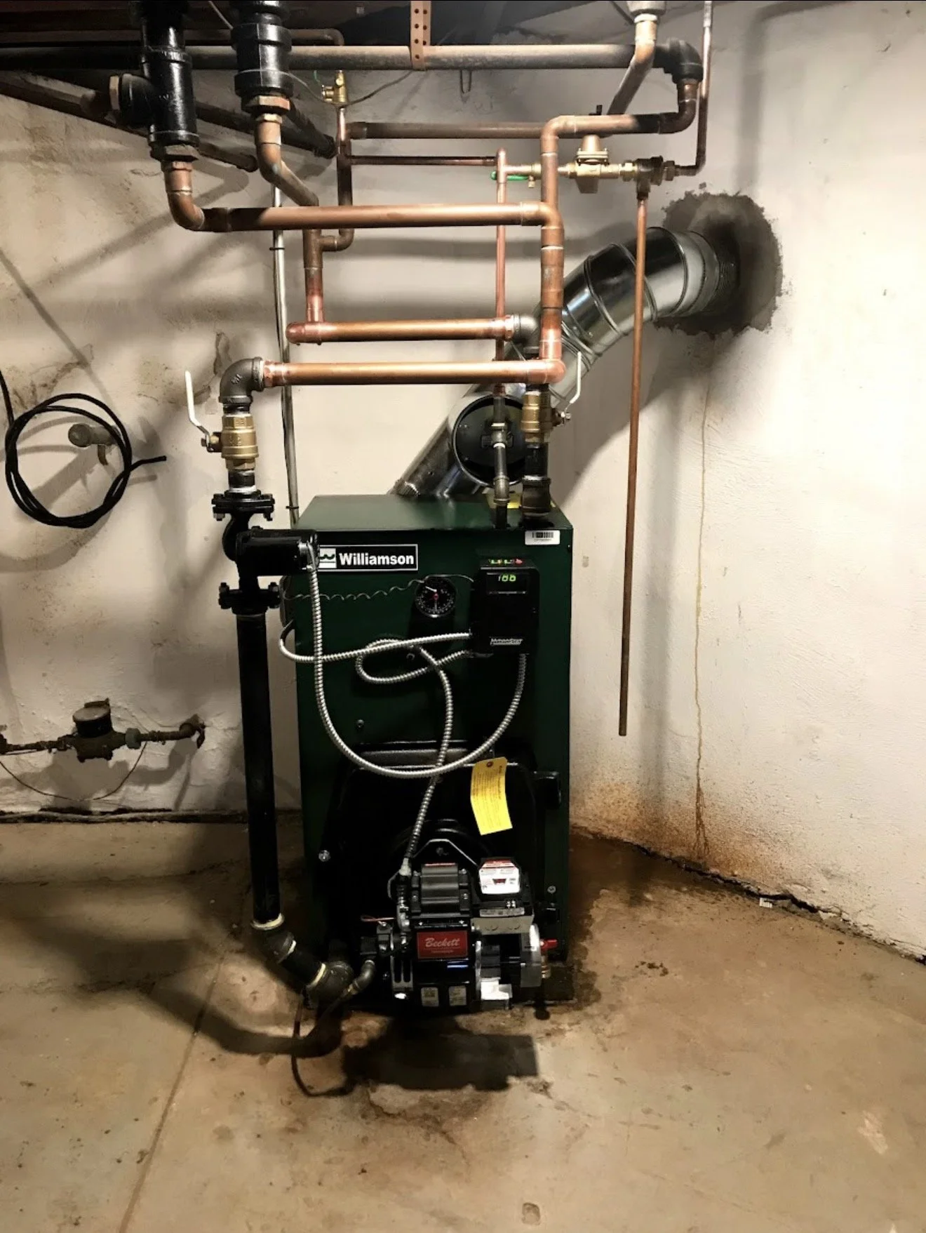 A green Williamson boiler unit installed in a basement or utility room, with copper pipes connected to it, and a vent pipe extending through the wall.