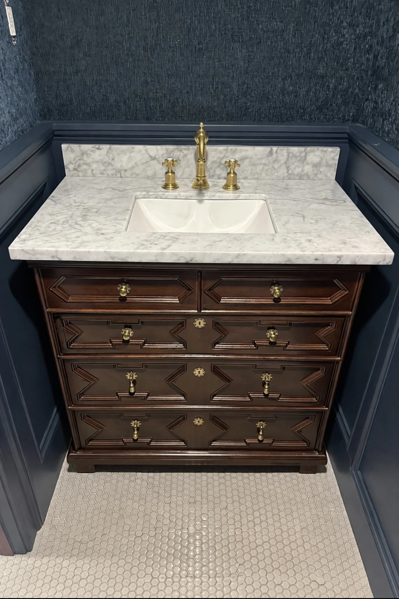 A vintage-style bathroom vanity with a marble countertop, gold fixtures, and a brown wooden cabinet with decorative carvings and gold knobs. The floor is covered with small white hexagonal tiles.