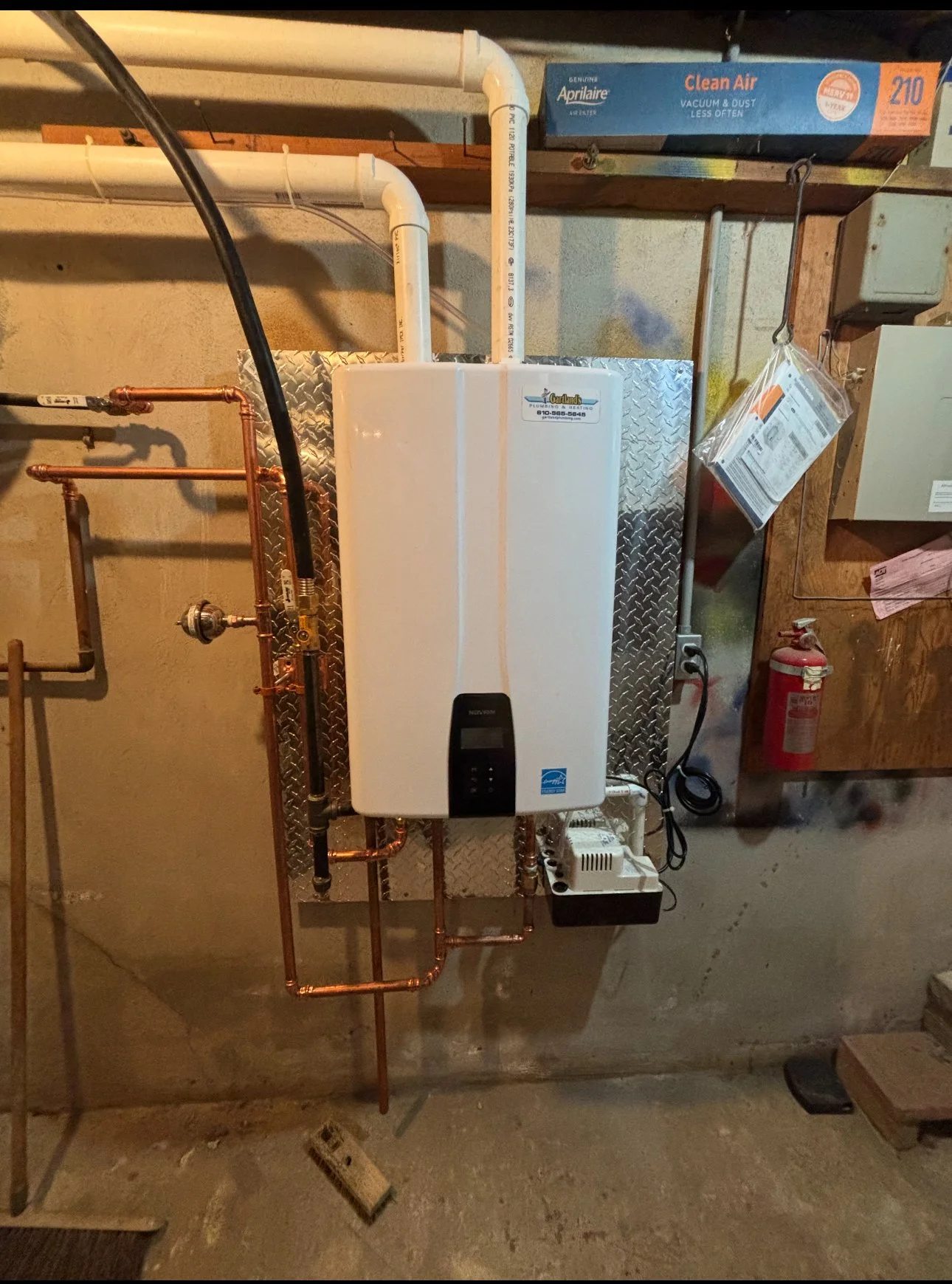 A white tankless water heater mounted on a concrete basement wall, with copper pipes and a white PVC vent pipe.