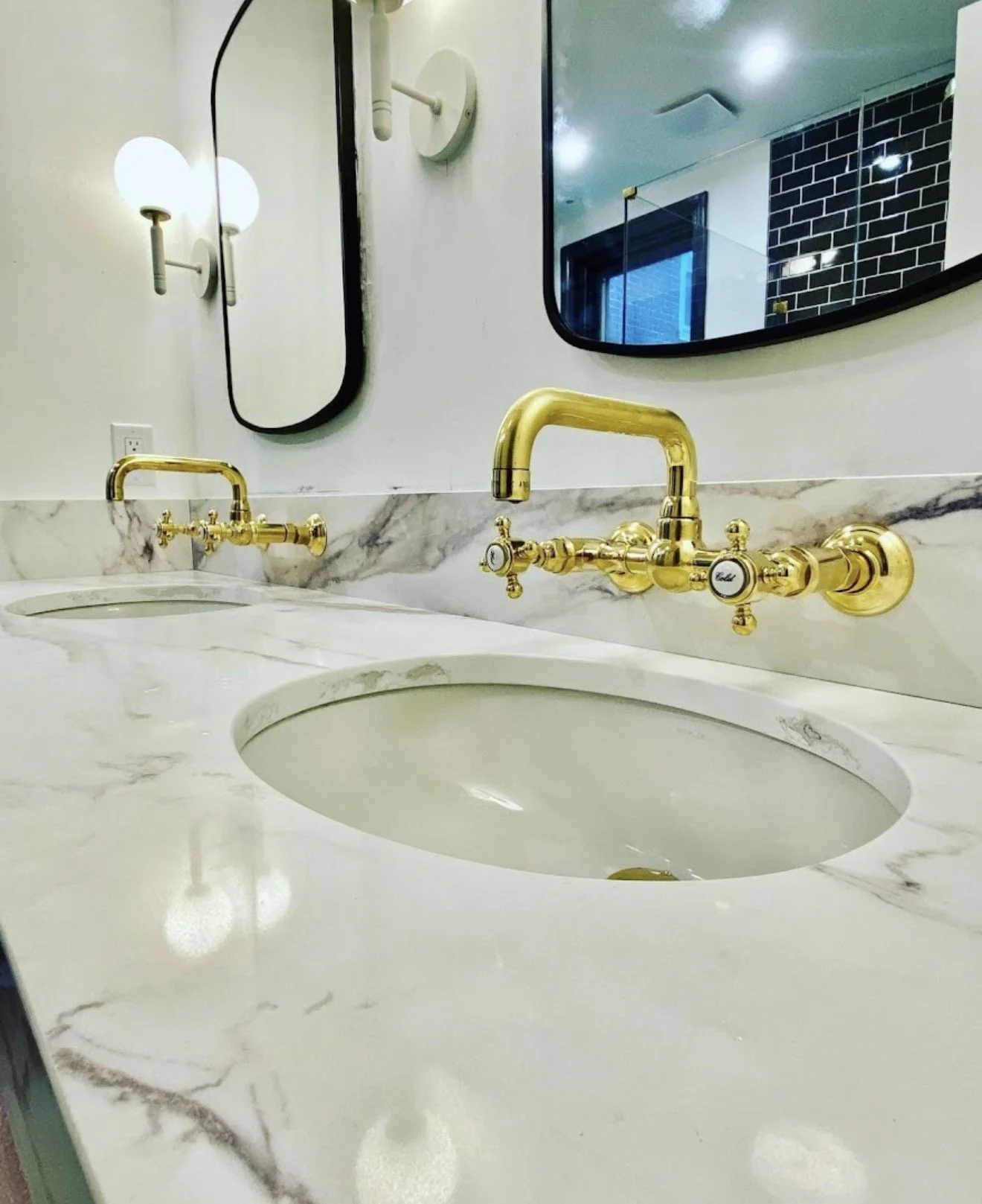 Close-up of a marble bathroom sink countertop with two gold faucets and oval sinks, with a large mirror and wall-mounted light fixtures in the background.