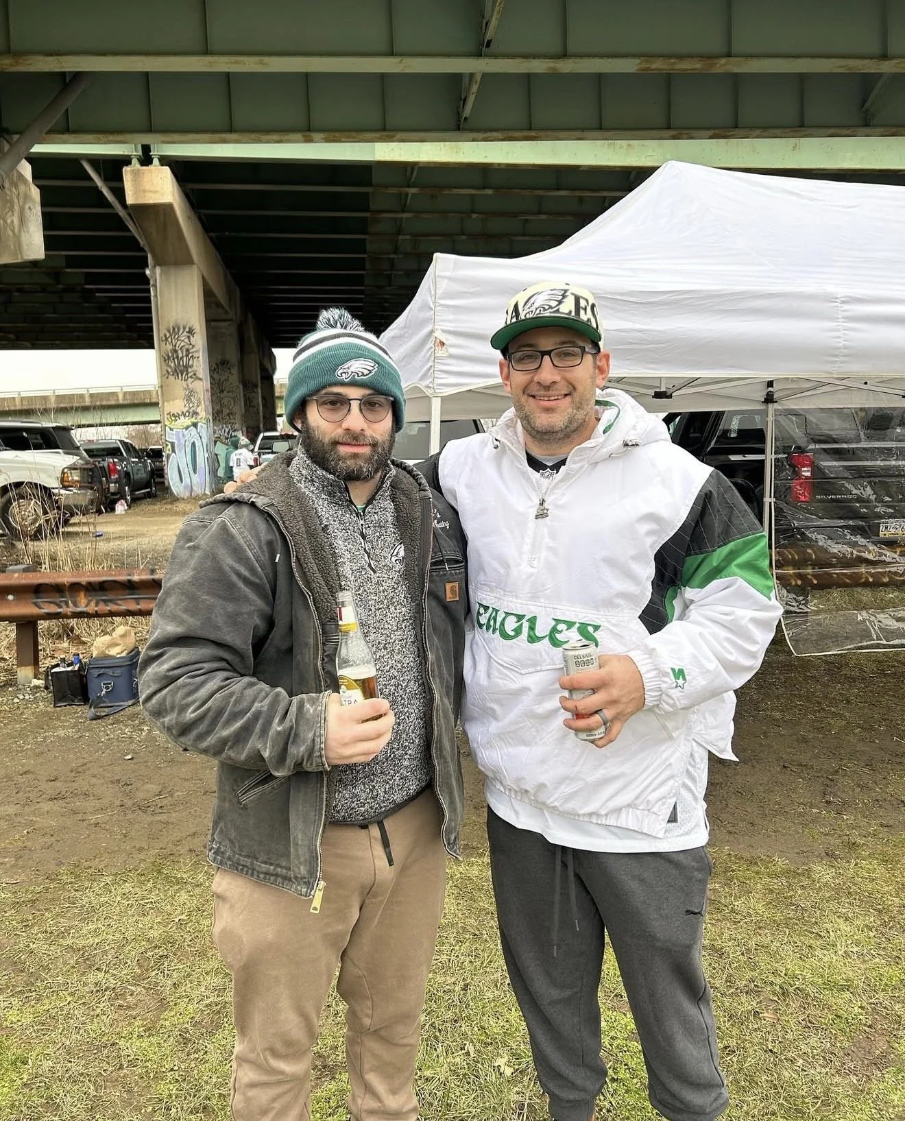 Two men standing under a bridge with tents and parked cars in the background, holding drinks, one wearing a Philadelphia Eagles hat and the other wearing glasses and a Eagles cap.