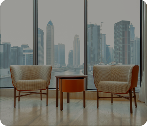 Two beige armchairs with wooden legs facing each other and a small round wooden table in between, set against large window panes showing a city skyline with tall buildings.