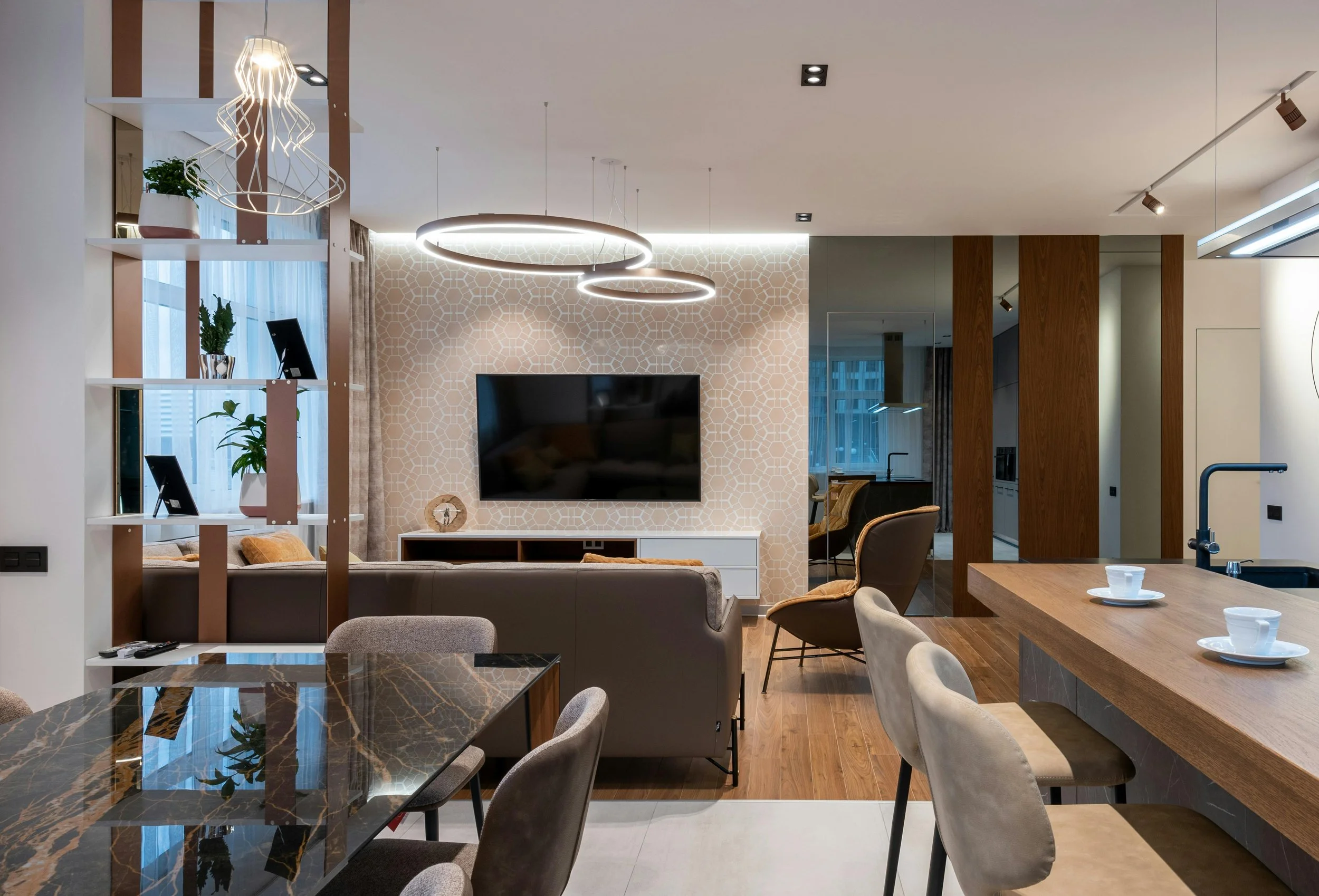 A modern open-plan living and dining area with a dining table, chairs, a TV mounted on a patterned accent wall, and various decorative elements including plants and lighting fixtures.