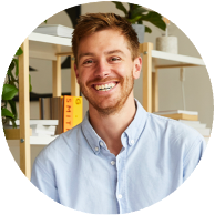 Smiling man with red hair and beard in an office, wearing a light blue button-up shirt, with shelves and office supplies in the background.