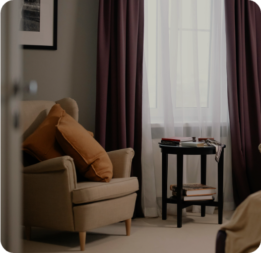 A cozy living room with a cream armchair with brown pillows, a small black side table with books, and purple curtains covering a window.