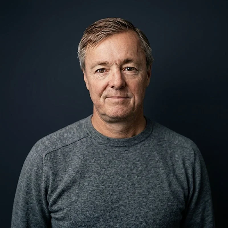 En medelålders man med grått hår, iklädd en grå tröja, tittar mot kameran mot en mörk bakgrund.