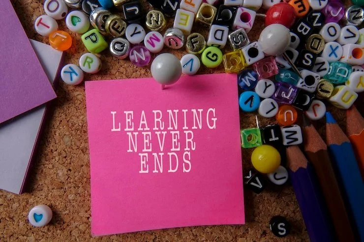 A pink sticky note with the phrase "Learning Never Ends" written in white letters, surrounded by colorful beads, markers, and paper on a corkboard surface.