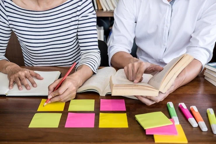 Two people are sitting at a wooden table with open books, sticky notes, and highlighters, discussing or planning together.