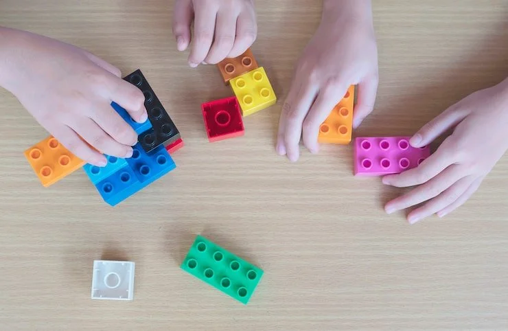 Two children playing with colorful plastic building blocks on a wooden surface.