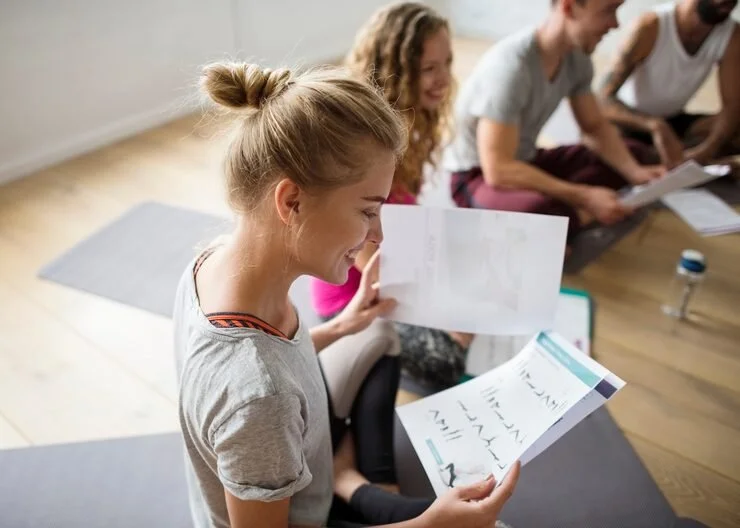 A group of four young people sitting on the floor, reading papers and smiling during a casual activity or class inside a room with wooden flooring.