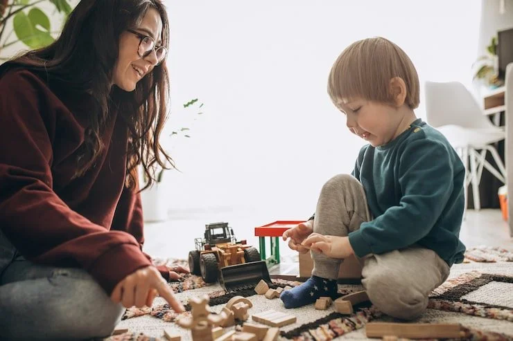 A woman and a young boy playing with wooden toys and a toy bulldozer on a rug indoors.
