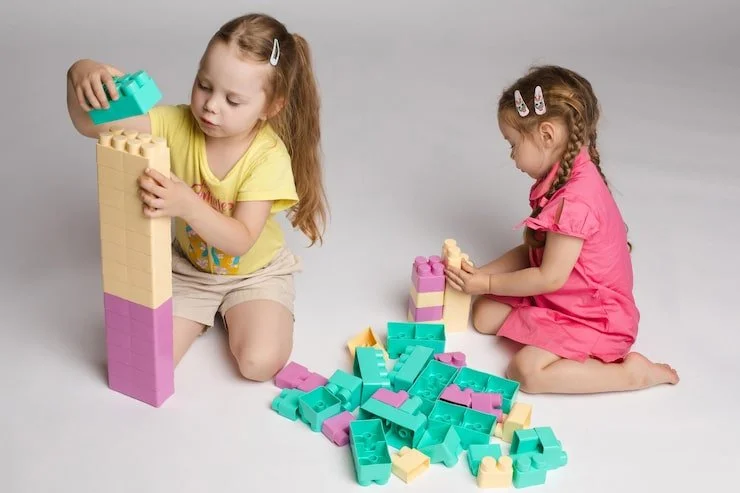 Two young girls playing with large colorful building blocks on a light gray background. One girl in a yellow shirt is stacking blocks, while the other girl in a pink dress is sitting and assembling blocks.