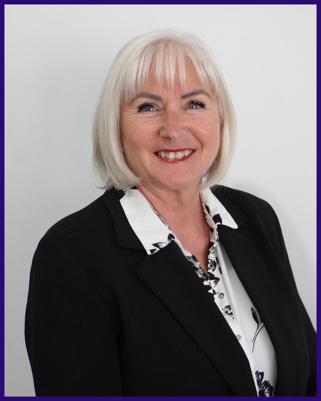 Headshot of a smiling middle-aged woman with blonde hair, wearing a black blazer and patterned white blouse, standing against a light background.
