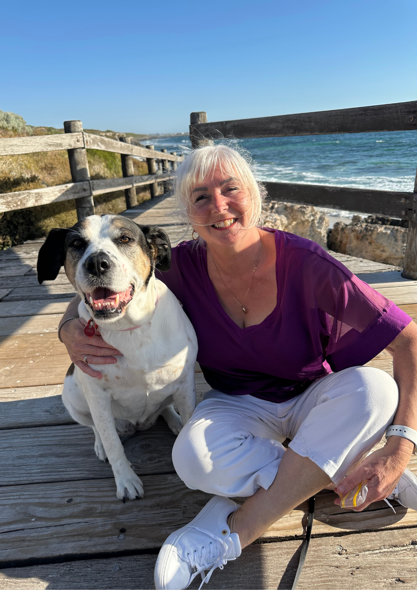 A smiling woman with white hair and a black and white dog sitting on a wooden pier by the ocean, with blue sky and waves in the background.
