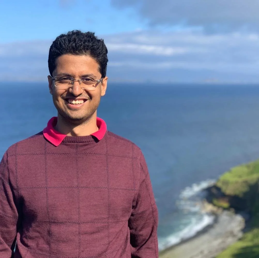 A smiling man with glasses standing outdoors near a coastline with the ocean and cliffs in the background.