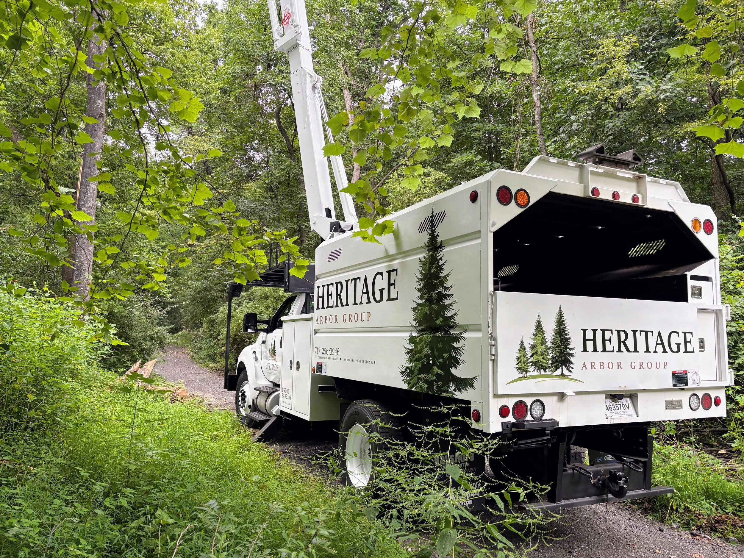 A white utility truck labeled "Heritage Arbor Group" parked on a wooded trail, with a hydraulic lift and a bucket for tree work, surrounded by green trees and foliage.