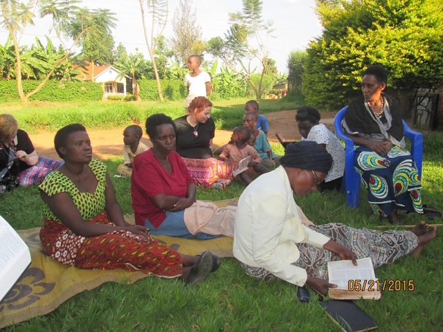 Group of women and children sitting outdoors on grass, some reading books, with trees and houses in the background, on a sunny day.