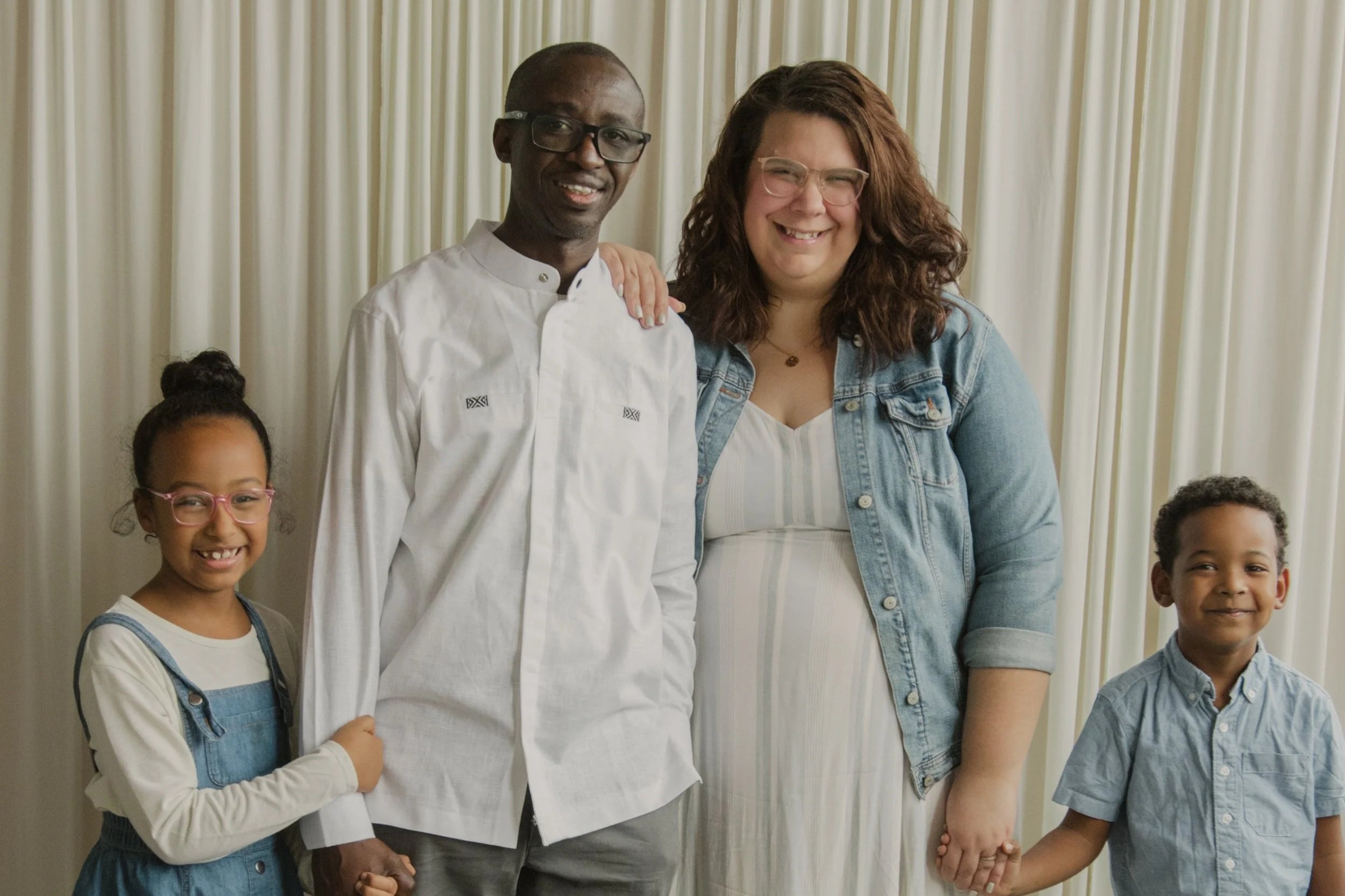 Group photo of a diverse family including two children and two adults, standing in front of cream-colored curtains, all smiling and holding hands.