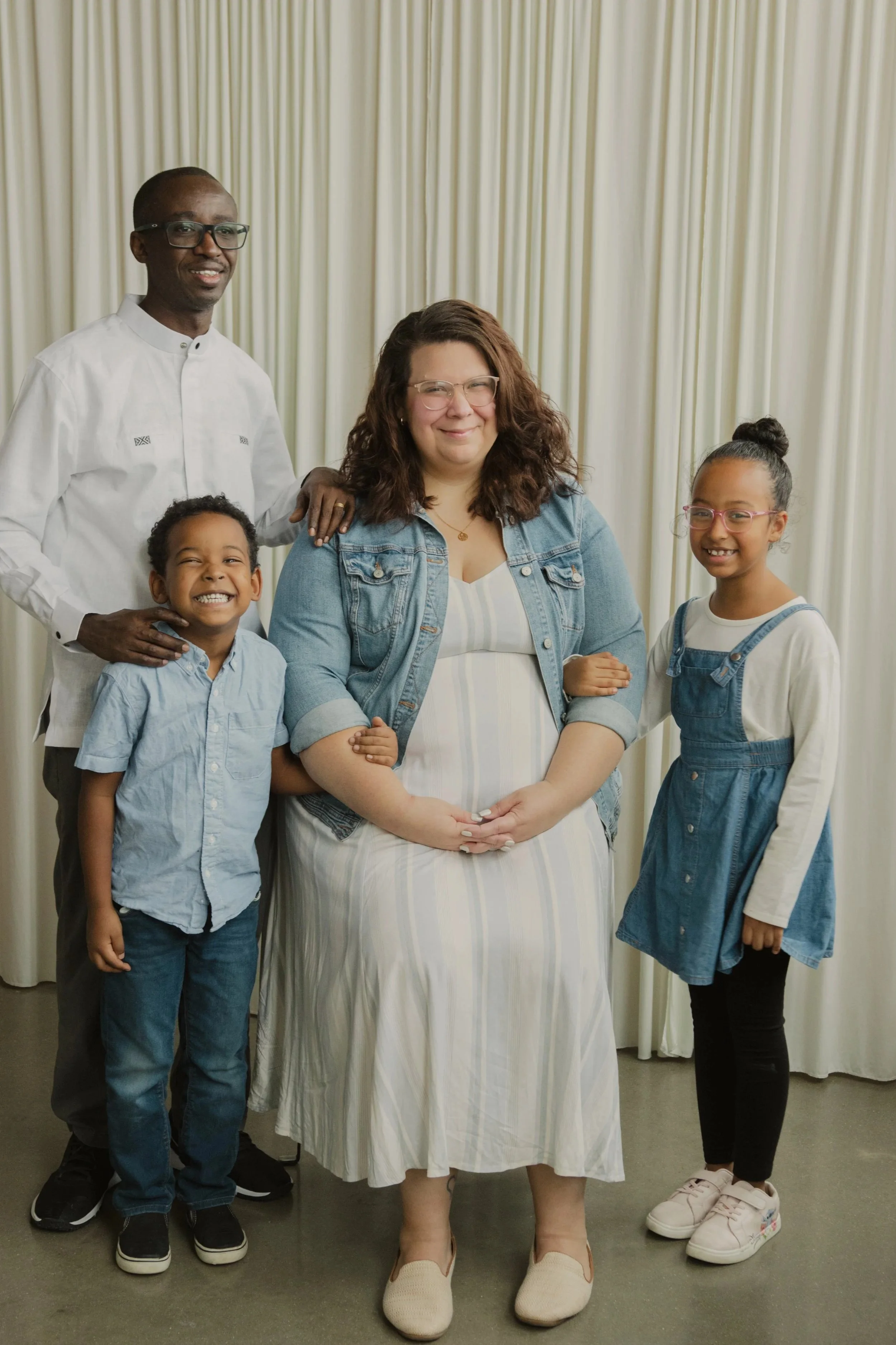 A diverse family group featuring two adults and two children, standing together indoors in front of cream-colored curtains, smiling at the camera.