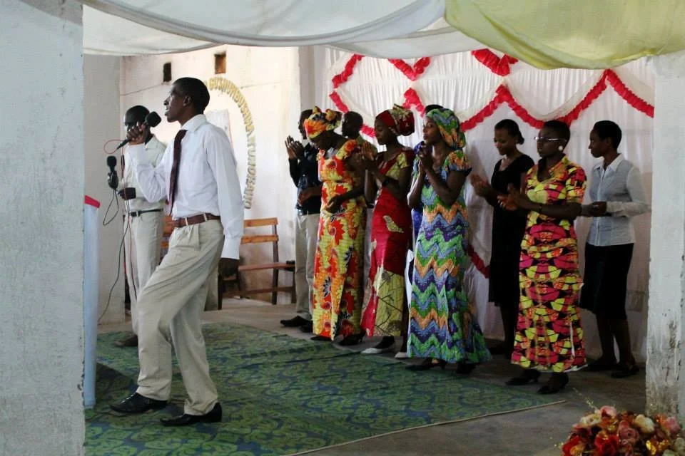 A man in a white shirt and beige pants speaking into a microphone on a stage with people clapping and standing behind him, decorated with white and red drapery, during a celebration or ceremony.