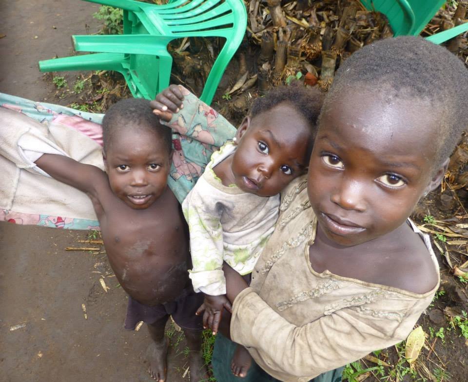 Three young children standing outdoors, two boys and a girl, with green chairs and fallen leaves in the background.