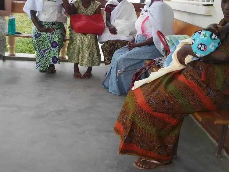 Group of women sitting and standing on benches in a waiting area, some with bags and children, in an outdoor setting.