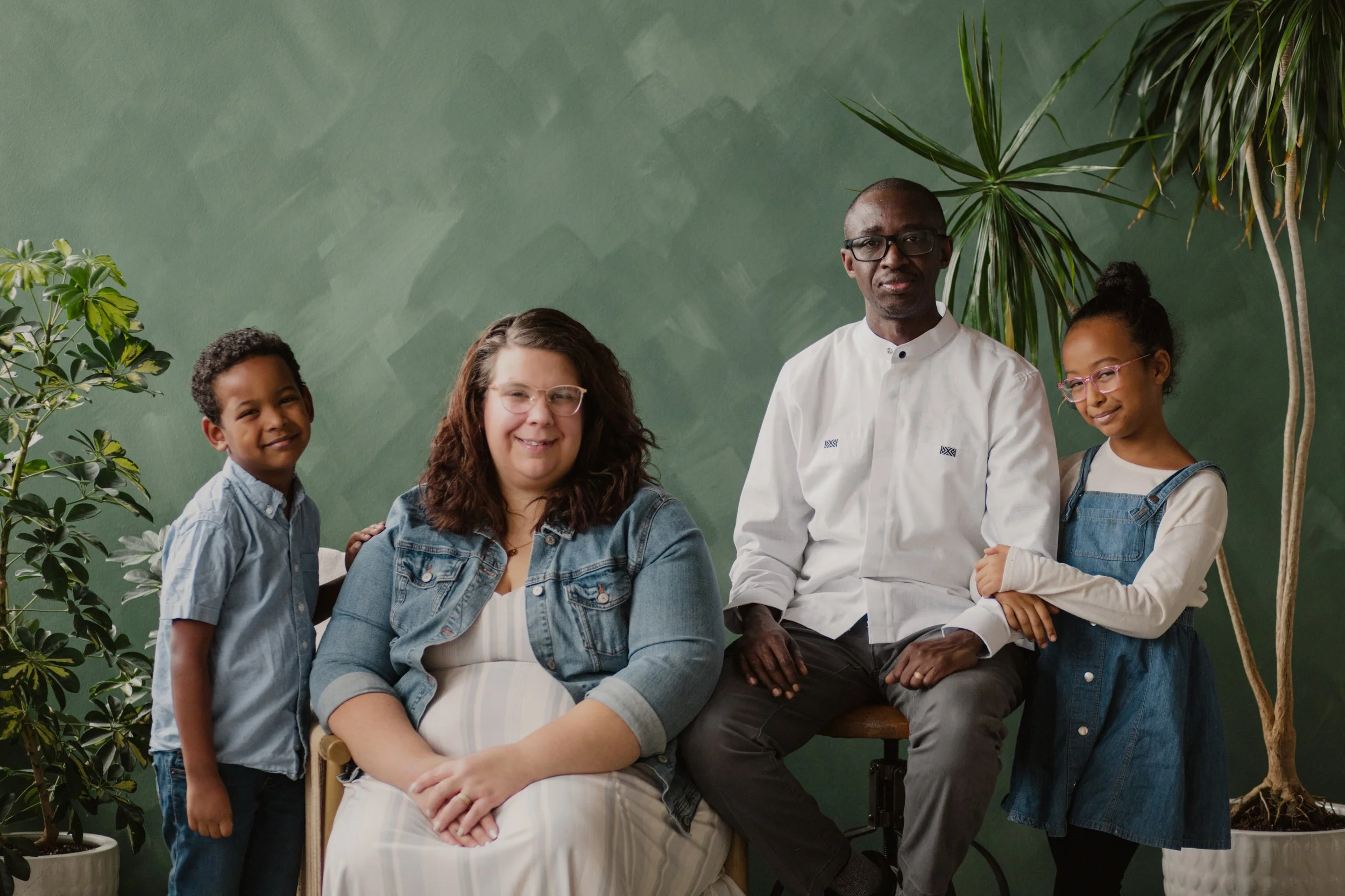 A diverse family portrait with two adults and two children standing and sitting in front of a green wall, surrounded by large indoor plants.